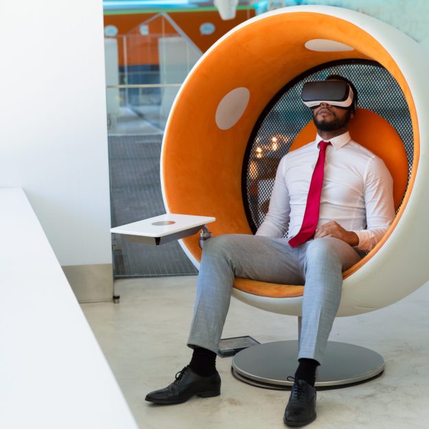 Peaceful businessman in VR headset enjoying virtual video. Man in office clothes and virtual reality goggles sitting in interactive chair and relaxing. Virtual content watching concept